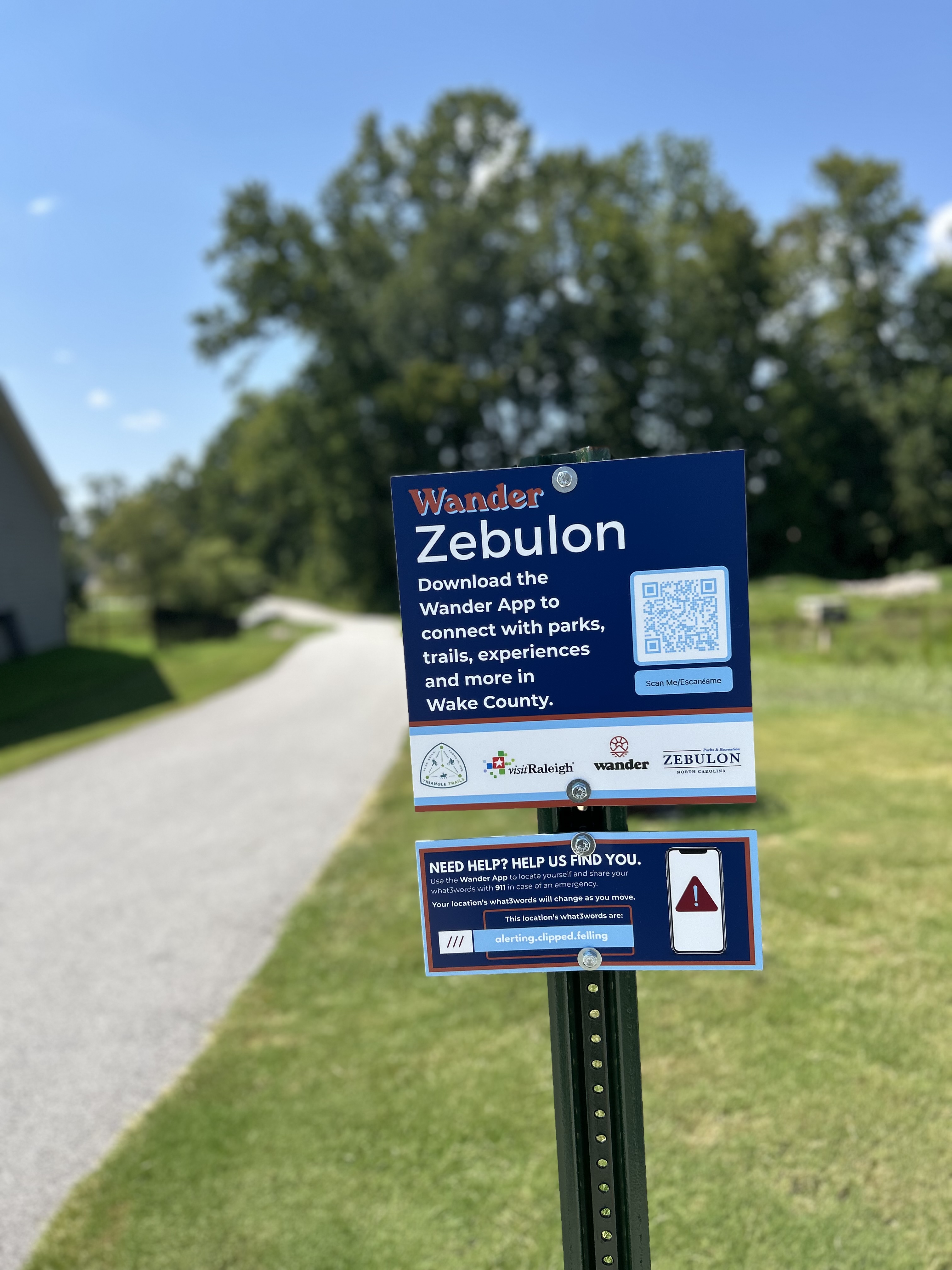 An installed blue Wander Zebulon sign beside a paved path, displaying a QR code and the partner logos.